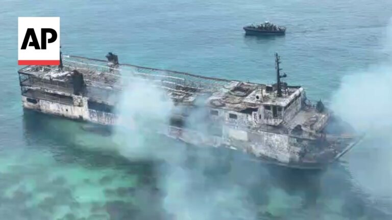 A large, rusted shipwreck floating in calm turquoise water with smoke rising from its deck.