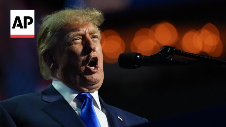 Man speaking into a microphone at a podium, with an AP logo in the corner and blurred orange lights in the background.