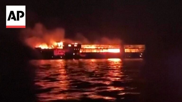 Night scene of a boat on dark water engulfed in flames with flames reflecting on the waves; AP logo top-left.