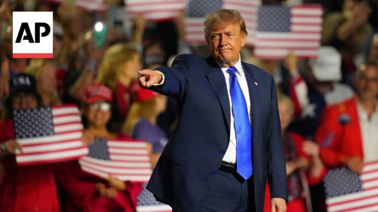A man in a dark suit and bright blue tie points toward the crowd at a rally, with American flags visible in the background and an AP logo in the top-left corner.