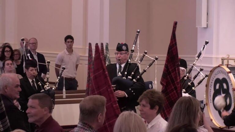 Bagpipers in uniform perform in a hall while seated audience watches a formal event; plaid banners nearby and a large drum visible on the right.