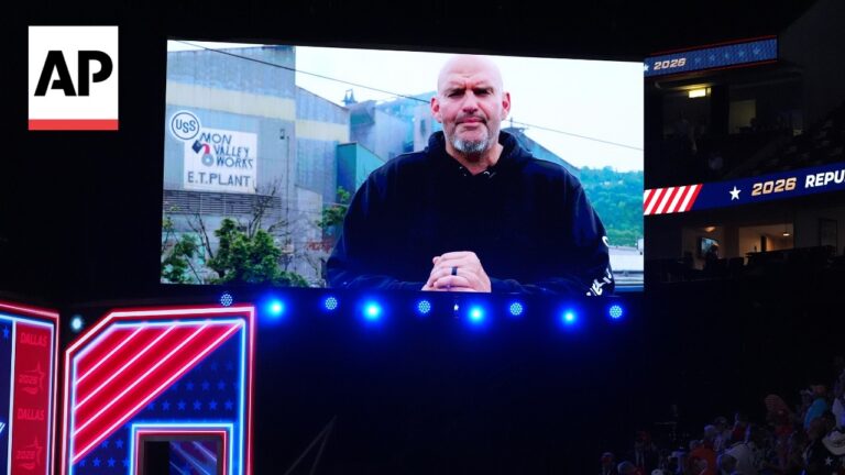 Large outdoor stage with a giant screen showing a man in a black hoodie speaking to an audience; AP logo in the top left and neon red-white-blue stage design on the left.