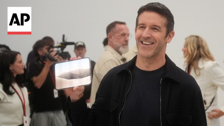 Smiling man in a dark jacket holds a smartphone displaying a desert landscape, AP logo in the top-left, photographers in the background.
