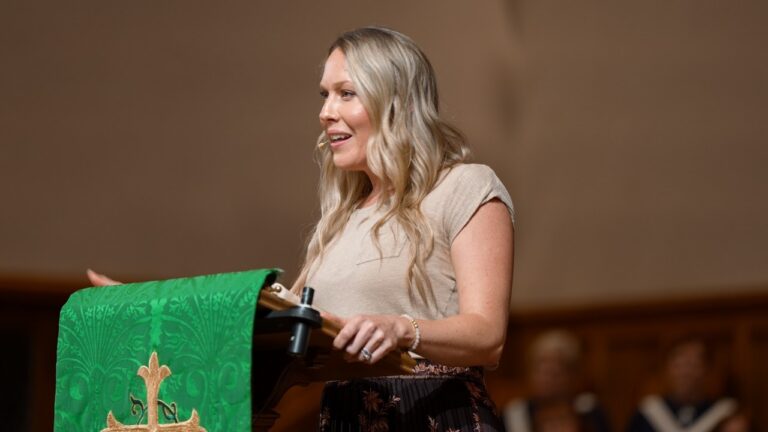 Woman speaking at a podium in a church, green lectern cloth with gold cross, wearing a beige top and bracelet