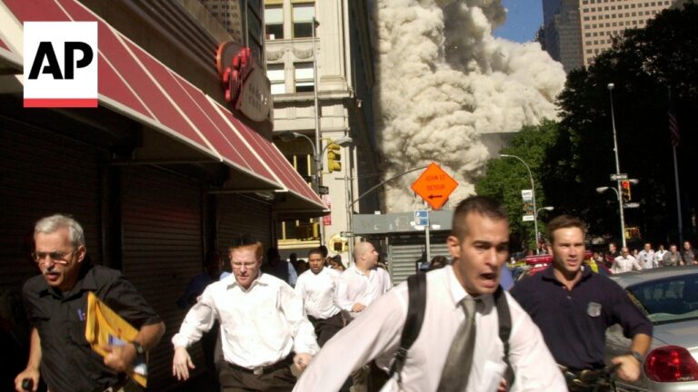 Group of people in business attire running down a city street as a large plume of smoke rises from behind a building; AP logo is visible in the top-left corner and an orange detour sign is in the background.
