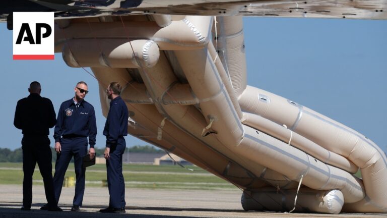 AP News logo in top-left; three men in navy uniforms stand on a tarmac beside a large beige inflatable aircraft emergency slide deflated and partially under a wing.