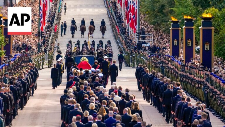 State funeral procession with rows of uniformed soldiers and a flag-draped coffin on a hearse ahead, crowds along the avenue, AP watermark in the corner.