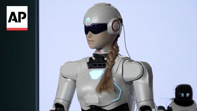Silver humanoid robot with braided hair, wearing visor and headset, on stage with AP logo.