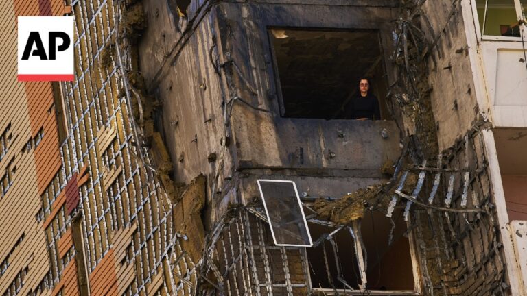 Wide-angle shot of a heavily damaged high-rise with exposed concrete and debris, a person in dark clothing stands in a broken upper window looking out. AP watermark visible on left.