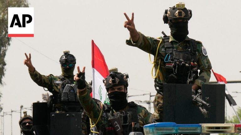 Masked soldiers in camouflage gear atop an armored vehicle, flashing victory/peace signs with an Iraqi flag in the background (AP photo).