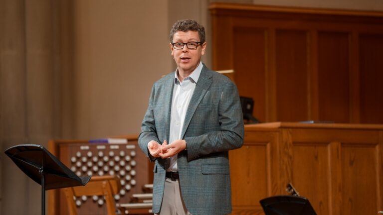 Male speaker in a grey blazer presenting on stage in a wooden-panel auditorium, gesturing with hands
