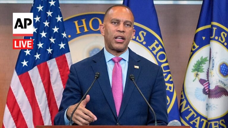 Man in a blue suit and pink tie speaks at a podium, with American flags and a government seal in the background, AP Live badge visible.