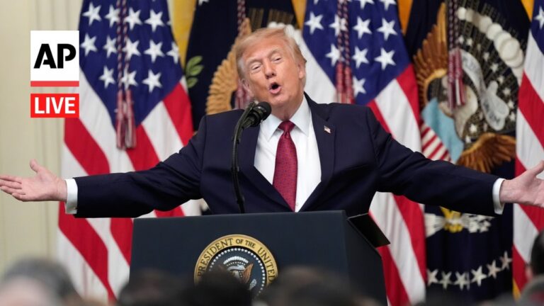Man in a dark suit and red tie speaks from a podium with the presidential seal, arms outstretched, at a live AP event.