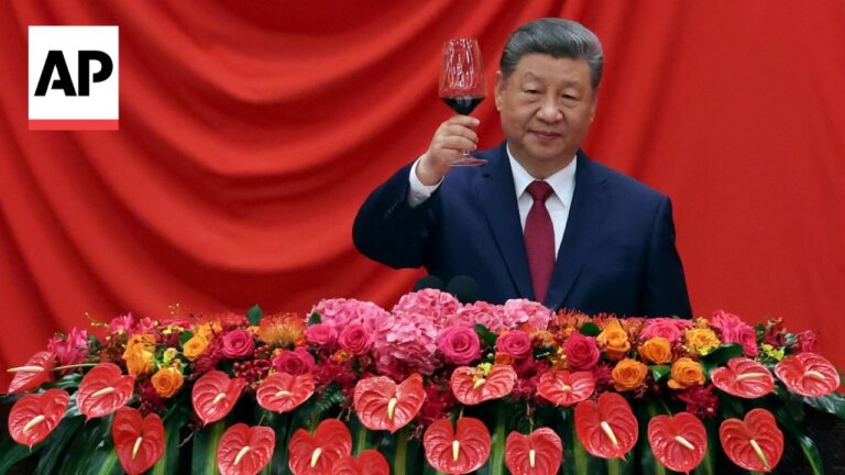 Man in a dark suit raises a glass of red wine at a podium with red flowers and an AP logo in the corner.