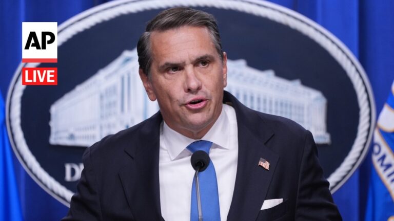 A suited man at a podium with a microphone giving a press conference, wearing a blue tie and an American flag pin, with AP Live logo in the background.