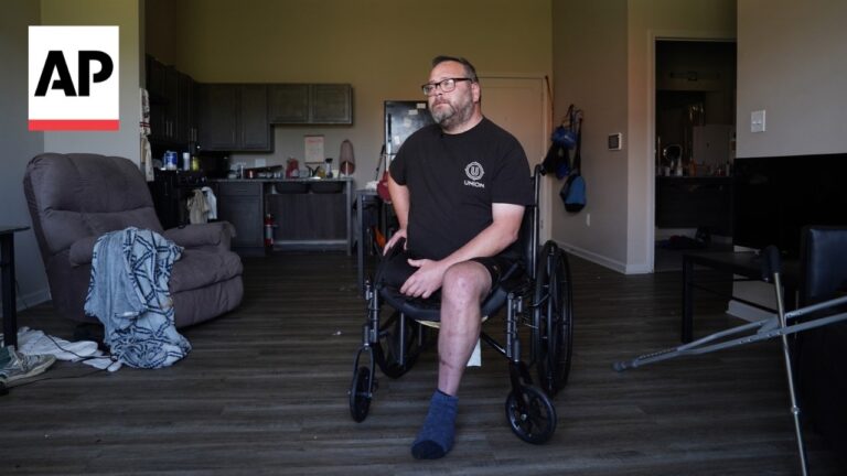 Man in a wheelchair sits in a living room with a kitchen in the background; AP logo in the top left corner.