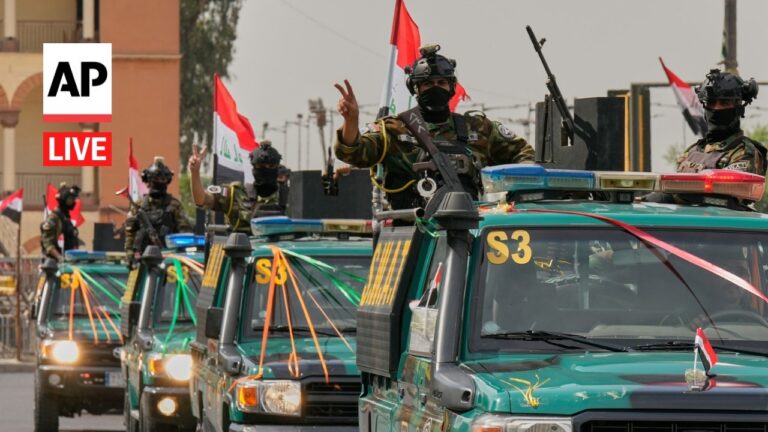 AP Live: soldiers wave from armored vehicles in a military parade with Iraqi flags overhead on a city street.