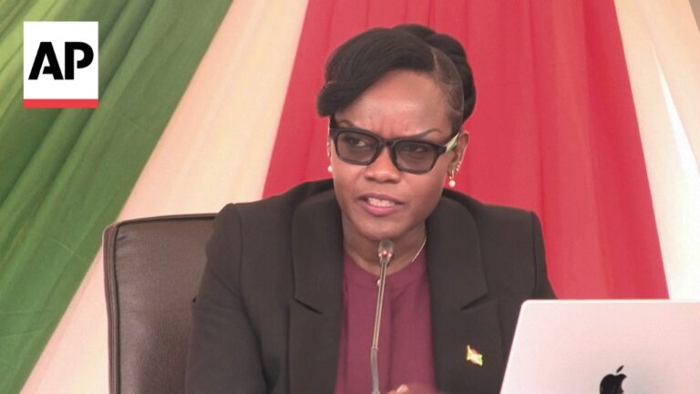 Professional woman in a black blazer speaks at a microphone during a press conference, AP logo visible in the corner.