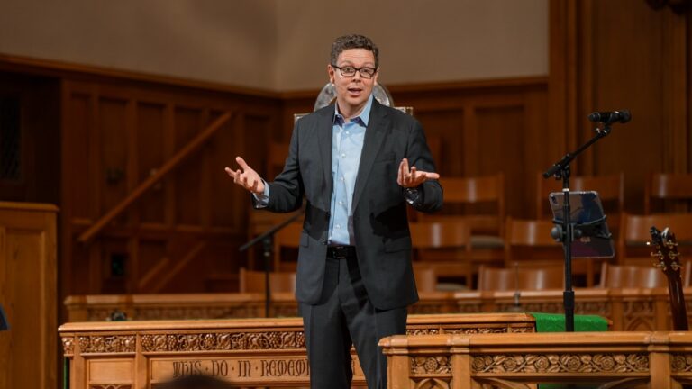 Man in a dark suit and glasses speaks from a wooden podium on a stage, gesturing with hands during a talk or sermon.