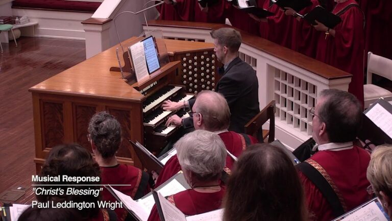 Choir in red robes singing in a church with an organist at a wooden pipe organ; on-screen caption reads Musical Response 'Christ's Blessing' by Paul Leddington Wright.