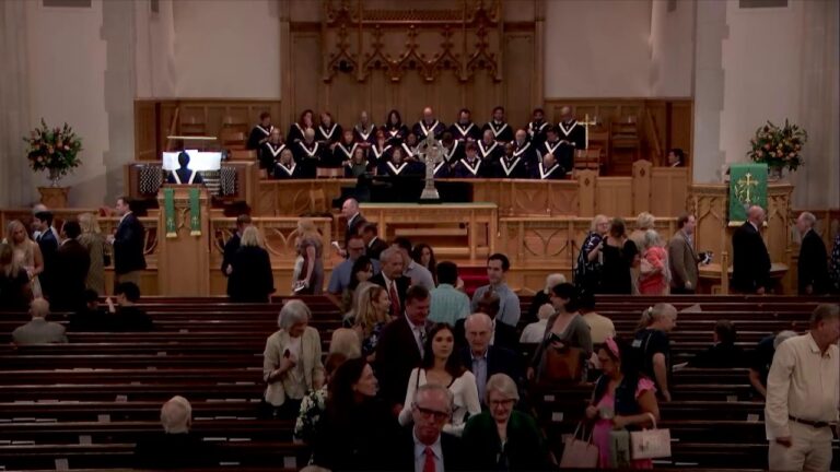 Inside a church sanctuary with a choir in black robes standing on a balcony behind an altar as people mingle in the aisle and pews below.