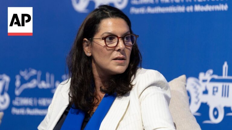 Woman with dark hair and red glasses seated on a panel, wearing a white blazer and blue scarf, against a blue backdrop with logos including AP in the corner.