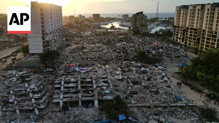 Aerial view of a coastal city in ruins: collapsed apartment blocks and rubble scattered across streets with people amid debris; AP logo overlaying the scene.