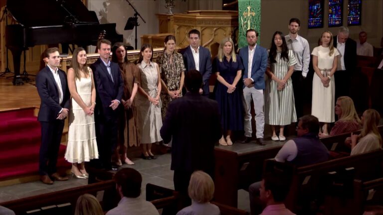 Conductor facing a line of formally dressed young adults on a church stage, ready to perform as seated audience watch from pews.