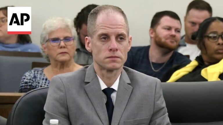 Man in gray suit and black tie sits in a courtroom, looking serious, with a seated audience behind him; AP logo top left.