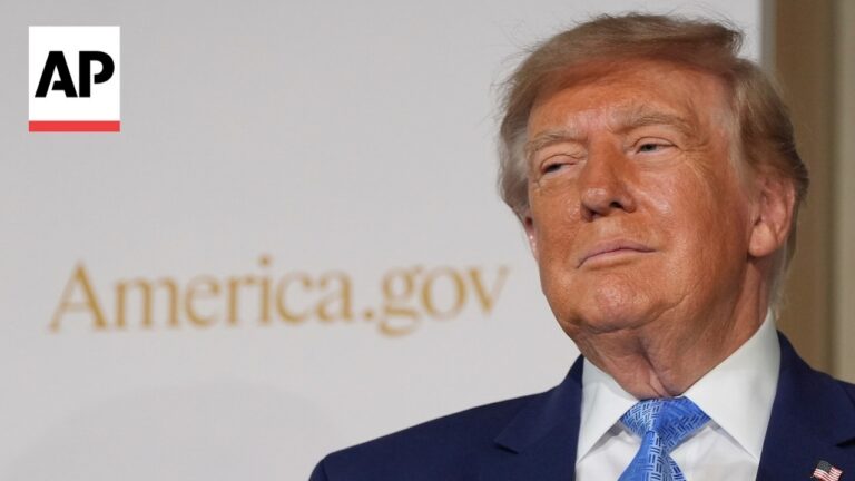 Close-up of a man in a navy suit and blue tie at a press event, with a blurred 'America.gov' sign and an AP logo in the top-left corner