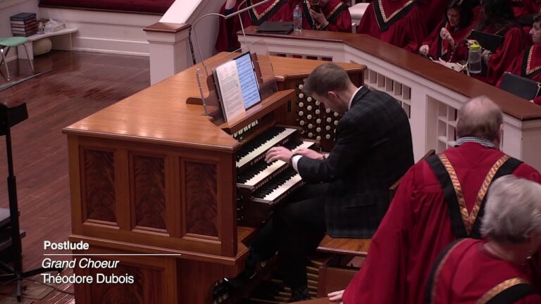 Organist in a dark suit playing a large wooden pipe organ as a choir in red robes looks on in a concert hall.