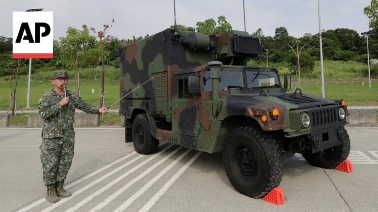 Military armored vehicle in camouflage with a soldier speaking into a microphone beside it, AP logo in the corner, outdoors on a paved lot.