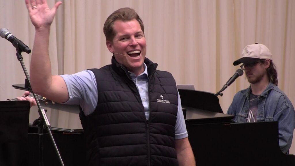 1792434580_maxresdefaultjpg - New Social Gospel Smiling man in a blue shirt and black vest waves to the audience, microphone headset on, with a pianist at a keyboard in the background.