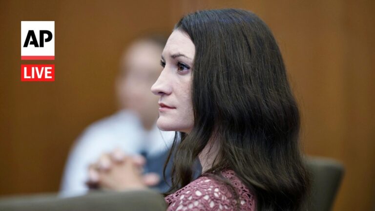 Profile of a woman with long dark hair wearing a pink lace top, seated in a courtroom; AP Live logo on the left.