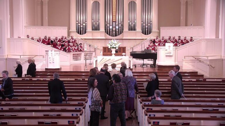 Interior of a church with a large organ behind a central altar and a choir on both balconies, people standing in the pews.