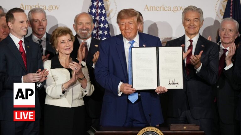 President Donald Trump holds an open, signed document at a podium during a ceremony, surrounded by smiling officials; AP Live watermark visible on left.