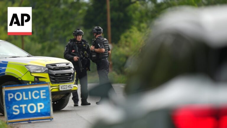 Two armed police officers stand beside a marked patrol SUV at a roadblock with a POLICE STOP sign nearby.
