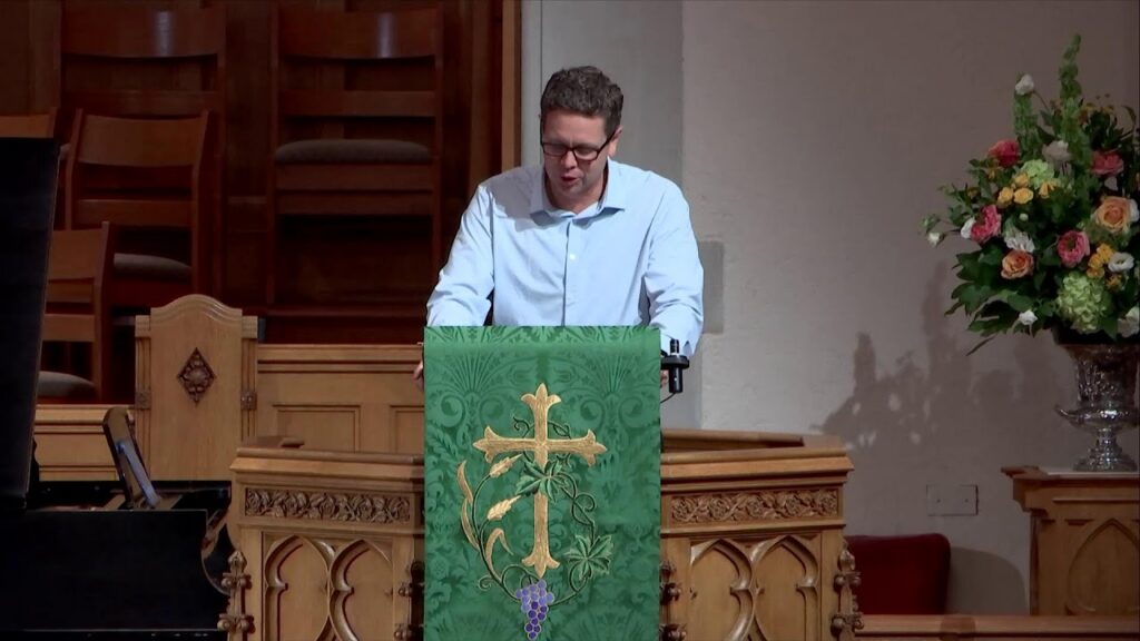 A man in a light blue shirt delivers a sermon at a church pulpit with a green cloth bearing a gold cross and grape motif, and a floral arrangement nearby.