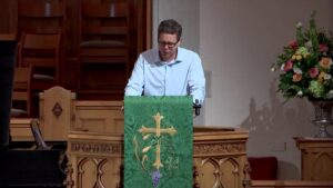 A man in a light blue shirt delivers a sermon at a church pulpit with a green cloth bearing a gold cross and grape motif, and a floral arrangement nearby.