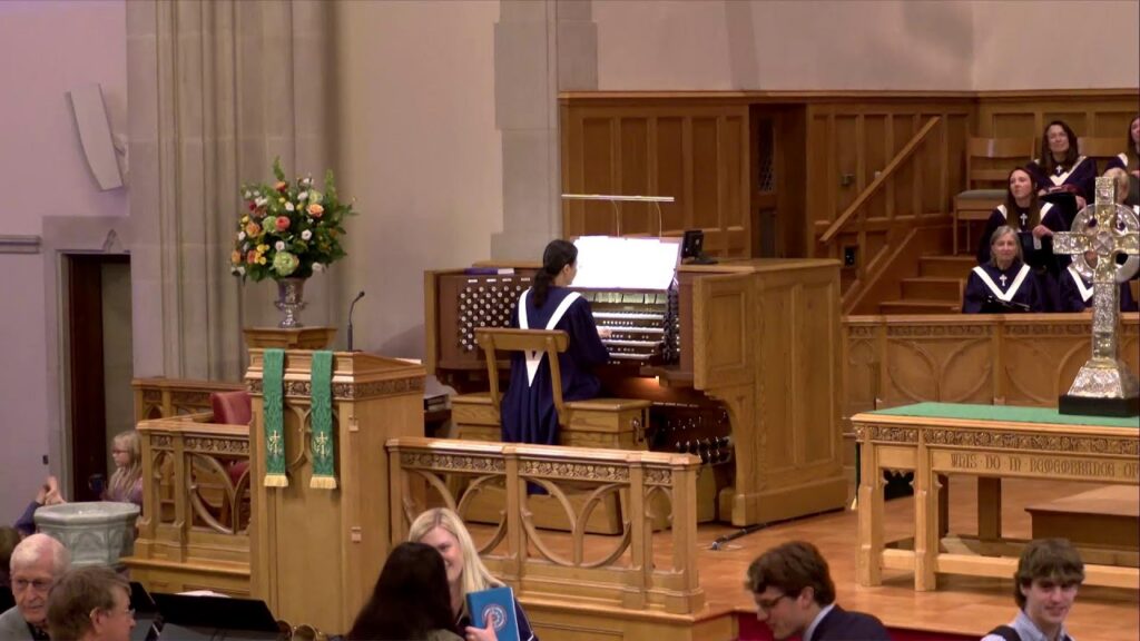 Organist in blue robe plays a pipe organ on a raised platform as a choir in purple robes sings nearby in a church sanctuary with wooden furnishings and a floral arrangement nearby.