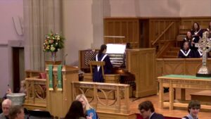 Organist in blue robe plays a pipe organ on a raised platform as a choir in purple robes sings nearby in a church sanctuary with wooden furnishings and a floral arrangement nearby.