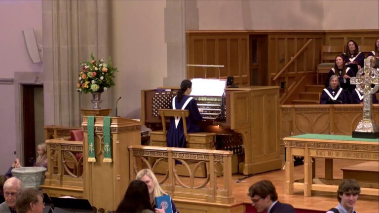 Organist in blue robe plays a pipe organ on a raised platform as a choir in purple robes sings nearby in a church sanctuary with wooden furnishings and a floral arrangement nearby.