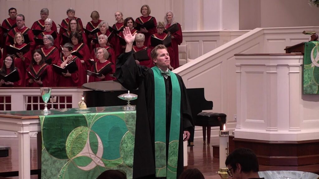 A choir in red robes stands behind a pastor at a green-aligned pulpit during a church service, the preacher raising his hand in blessing.
