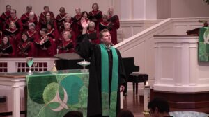 A choir in red robes stands behind a pastor at a green-aligned pulpit during a church service, the preacher raising his hand in blessing.