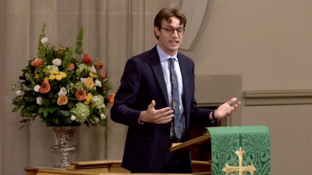 Man in a dark suit and tie speaking at a pulpit in a church, with a large floral arrangement beside him