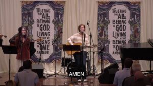 Worship band on stage with a guitarist and singer, banners behind reading 'Trusting that all belong to God, living like we belong to one another', audience seated