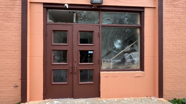 Brown double doors with small square windows, next to a large shattered pane in a peach brick storefront window.