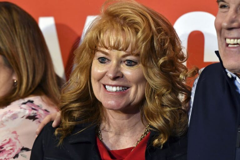 Smiling red-haired woman in a red blouse and dark jacket at a public event, with a man partially visible beside her.