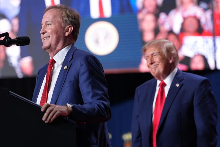 Two men in blue suits on a stage; one at a podium speaking into a microphone, the other smiling nearby with a blurred audience behind.