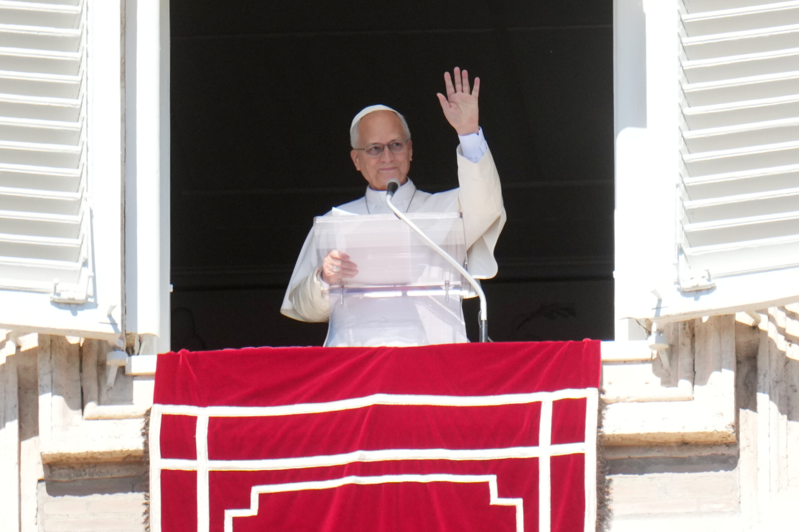 A Catholic cleric wearing white robes stands at a podium on a balcony, raising his right hand in a blessing gesture